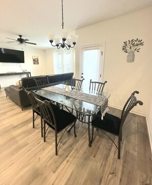 Un coin repas avec une table à plateau en verre entourée de chaises, un ventilateur de plafond et un téléviseur fixé au mur.