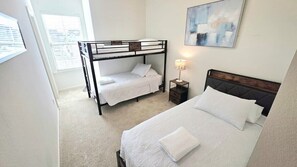 Une chambre avec un lit superposé, un lit simple, une table de chevet avec une lampe et un tableau accroché au mur.