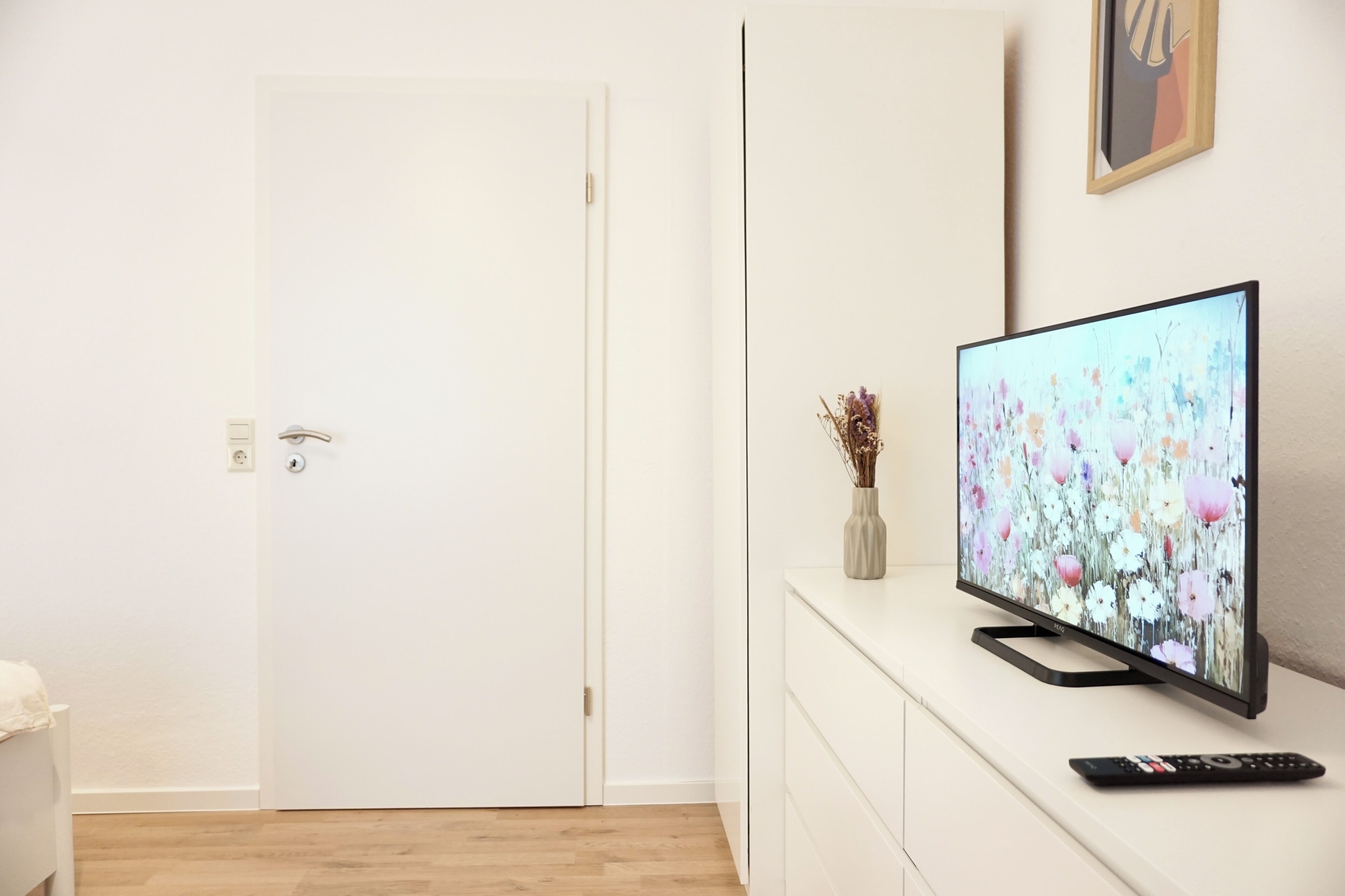 Habitació moderna amb una porta blanca, un televisió de pantalla plana sobre un moble blanc, i un comandament a distància sobre el moble.