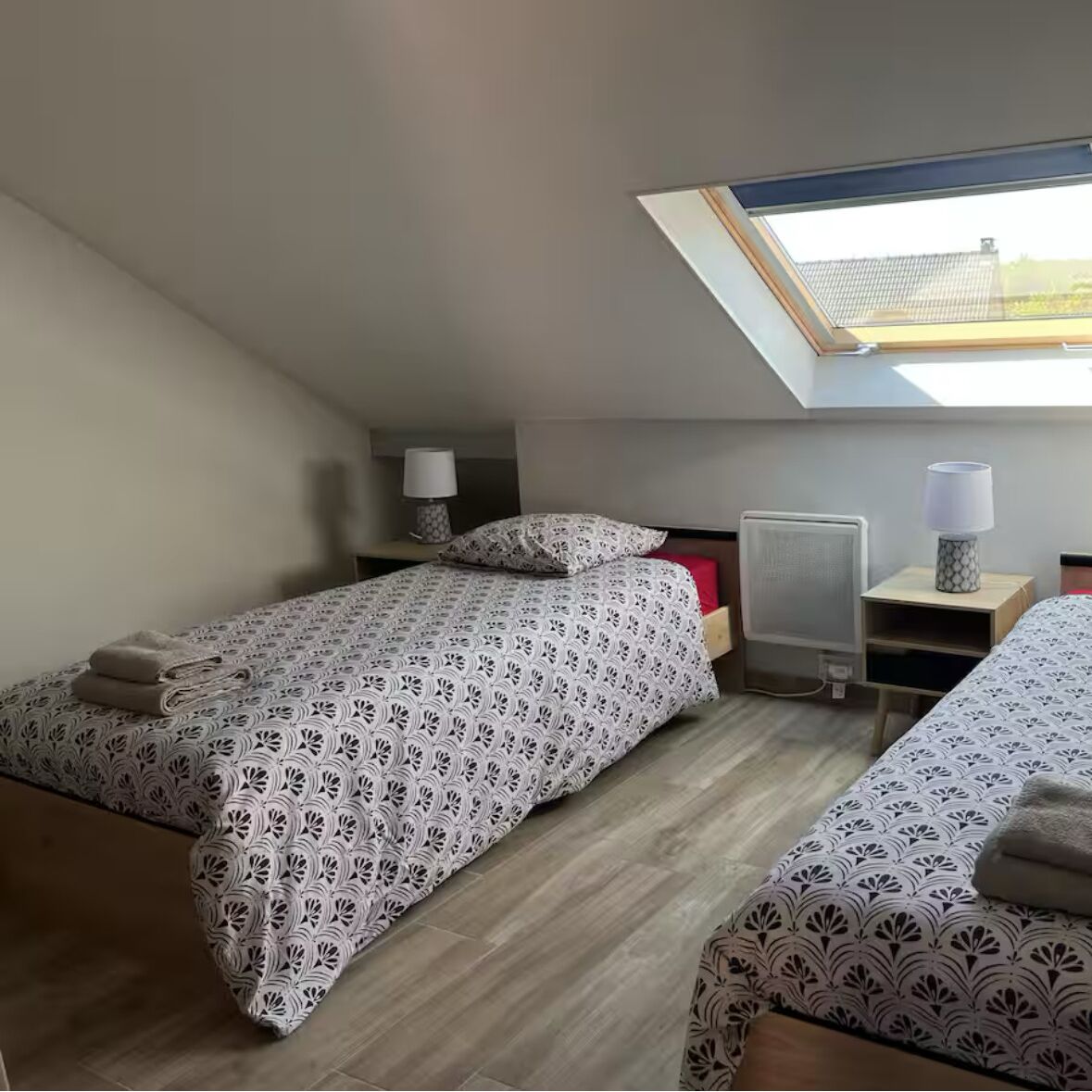 A small, modern bedroom with two beds, a skylight, a radiator, and a bedside table with a lamp.