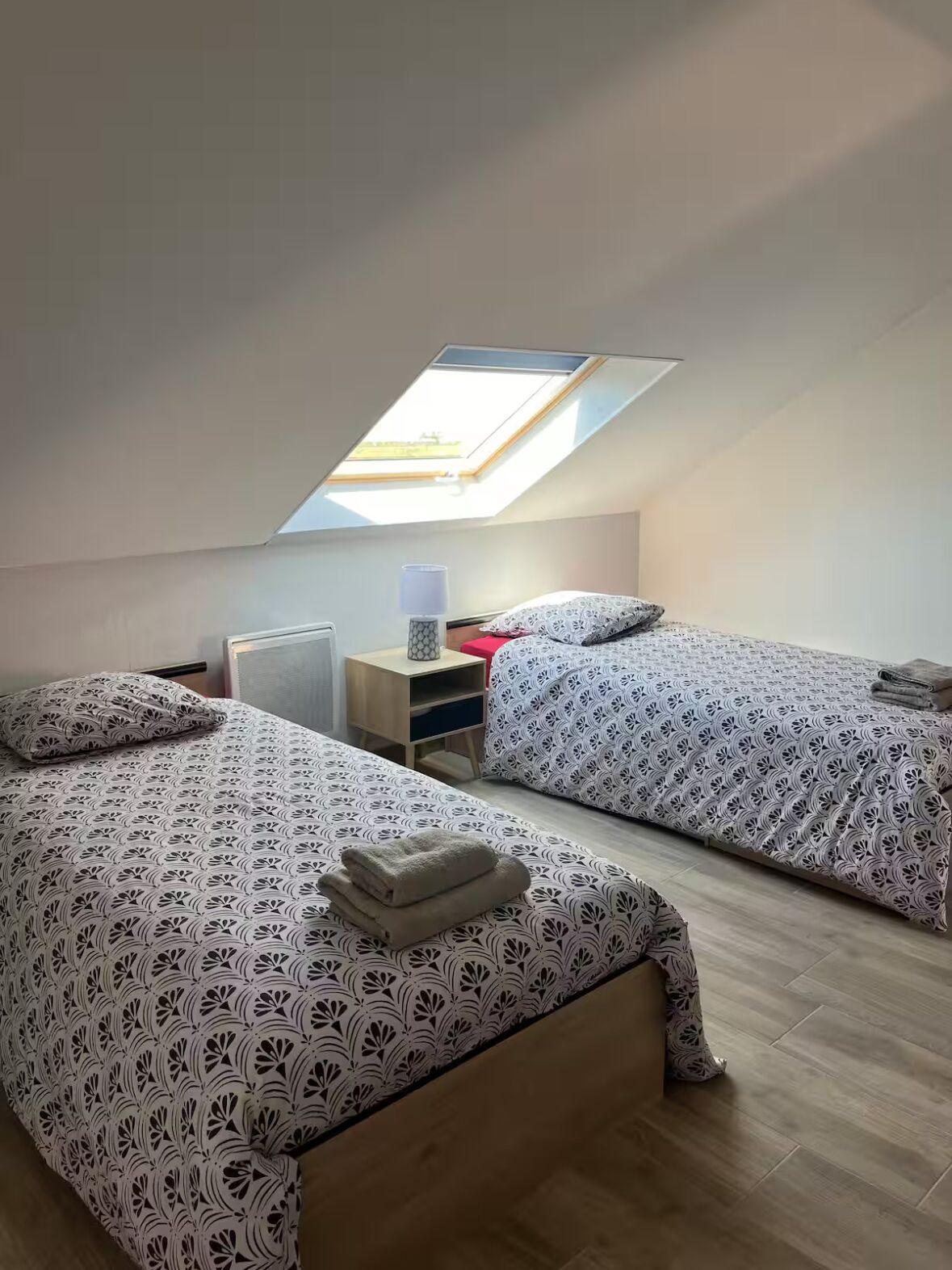 A dormitory room with two beds, a skylight, a bedside table with a lamp, and a radiator.