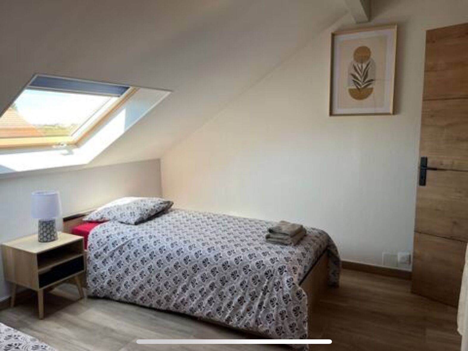 Un dormitorio con tragaluz, una cama con ropa de cama estampada, una mesita de noche con lámpara y una obra de arte enmarcada en la pared.