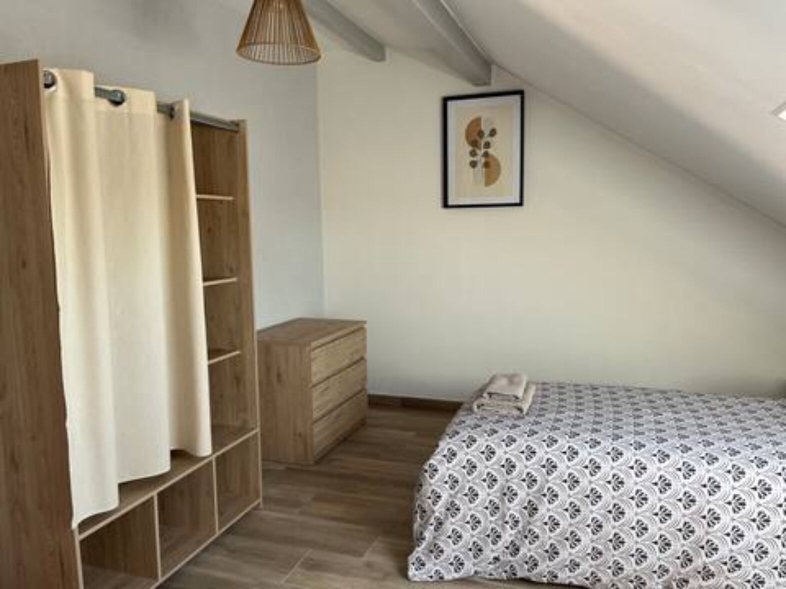 Ett sovrum med ett sluttande tak, en garderob i trä, ett litet sängbord, en säng med ett mönstrat överkast och ett inramat konstverk på väggen.
