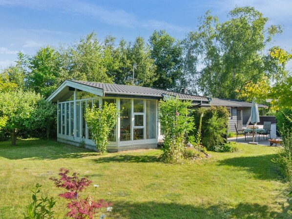 Een klein, laag huis met een glazen veranda, omgeven door een verzorgd gazon en diverse bomen.