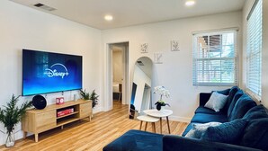 Un salon moderne avec un téléviseur à écran plat affichant le logo de Disney+, un canapé bleu, une petite table ronde et un meuble en bois orné d’une plante.