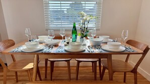 Une table à manger dressée avec de l’assiette blanche, des bols et des couverts, une bouteille verte et un vase rempli de fleurs blanches.