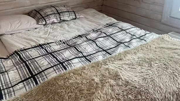 A cozy room with wooden walls, a bed with a striped bedspread, a fluffy rug, and a small shelf with a vase.