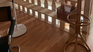 A room with wooden flooring, a standing fan, a wooden bench with a basket, and a view of the outside through a window.