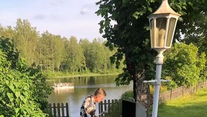A person tending to a portable grill near a lake, with a wooden fence and greenery in the background.