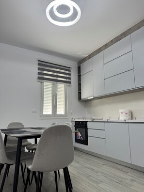A modern kitchen with a dining area, featuring a built-in oven, white cabinetry, and a window with blinds. - Charming 3-bedroom Villa in lovely Shkodër with AC (Shkodër)