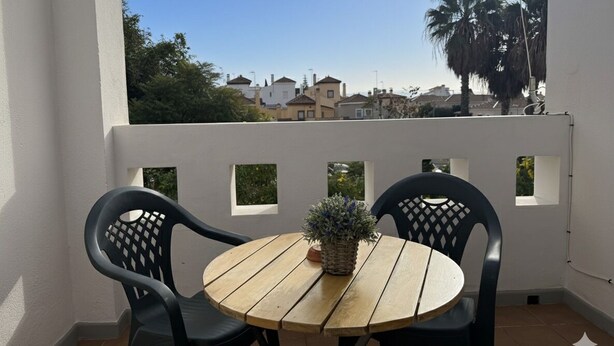 Ein Balkon mit zwei schwarzen Stühlen, einem Holztisch, einem Blumentopf und Blick auf Häuser und Palmen.