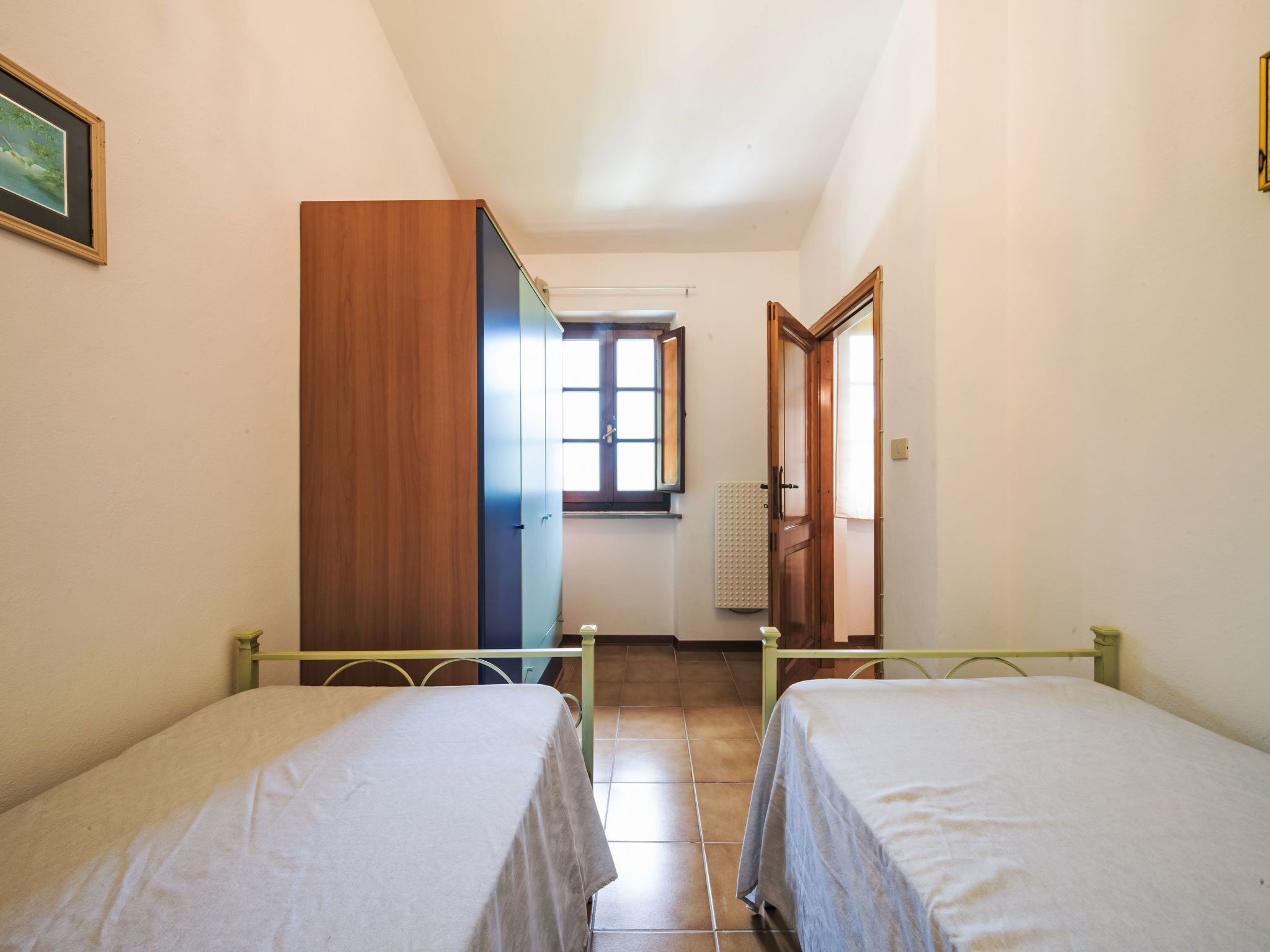 Une chambre avec deux lits, une armoire en bois, une fenêtre avec des stores et un radiateur.