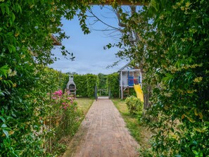 A brick pathway leading to a wooden playhouse with a slide, surrounded by lush greenery and flowering plants.