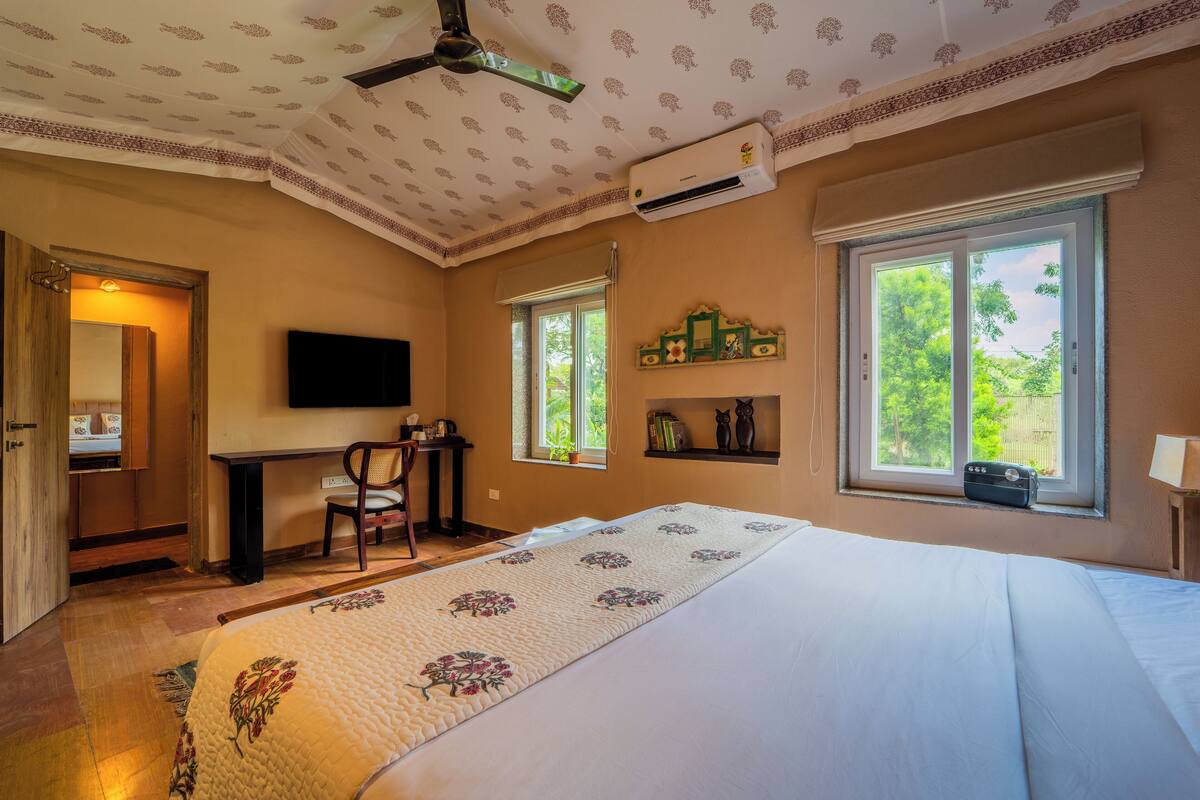 A bedroom with a large bed, a desk, a chair, a television, and a window with a view of greenery.
