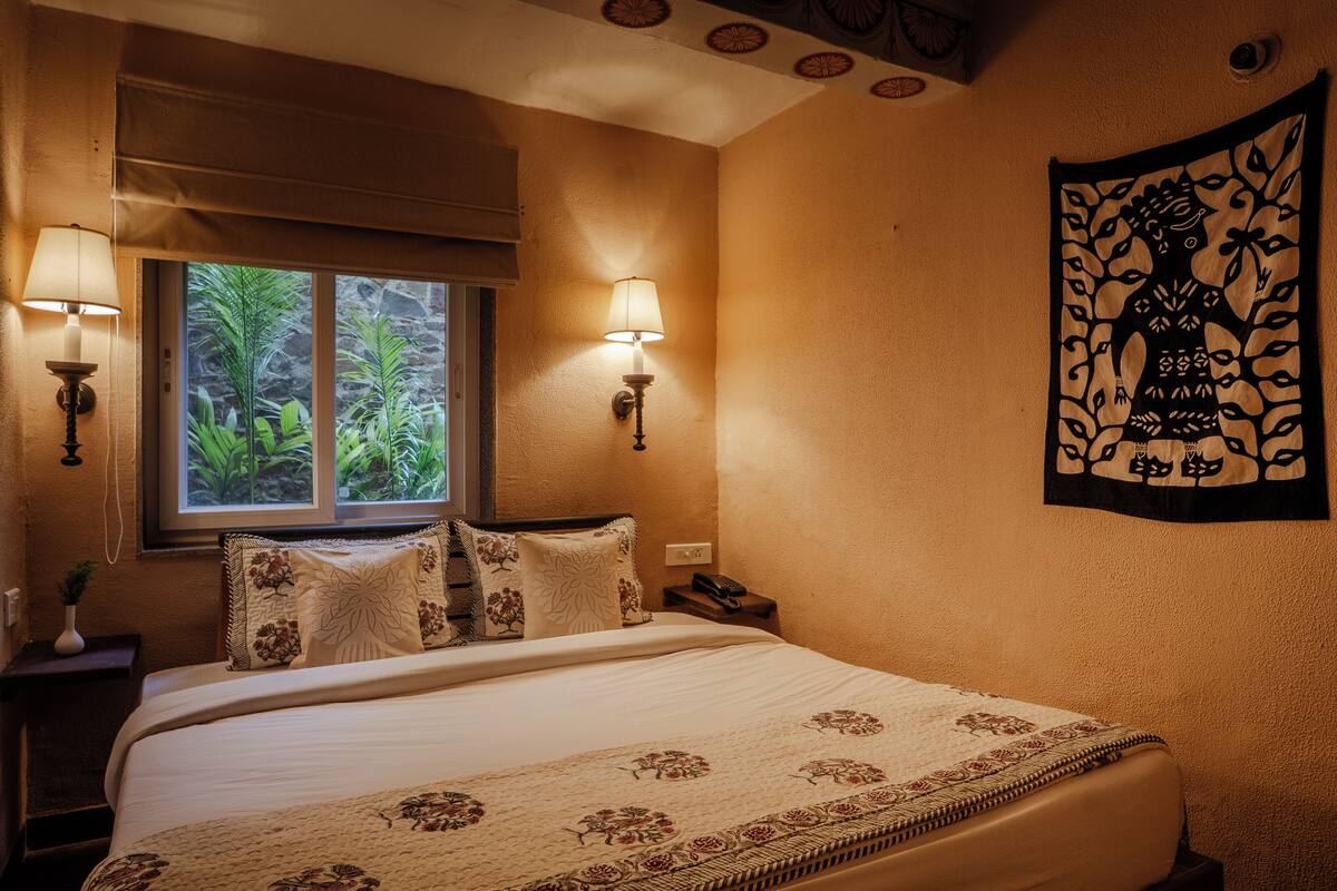 A hotel room with a large bed, two bedside lamps, a window with a view of greenery, and a tapestry on the wall.