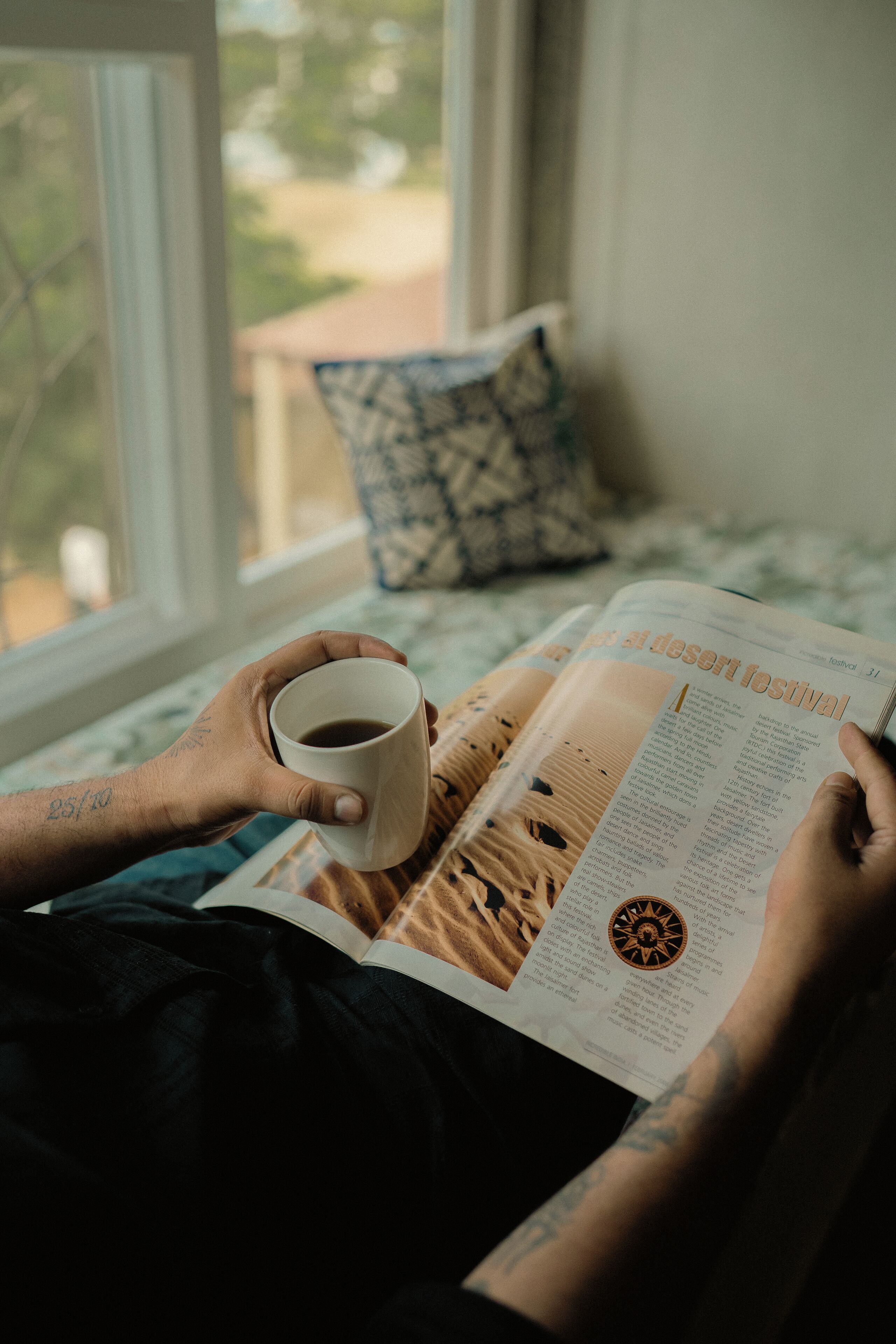 A person reading a magazine with a cup of coffee on a bed with a window view.