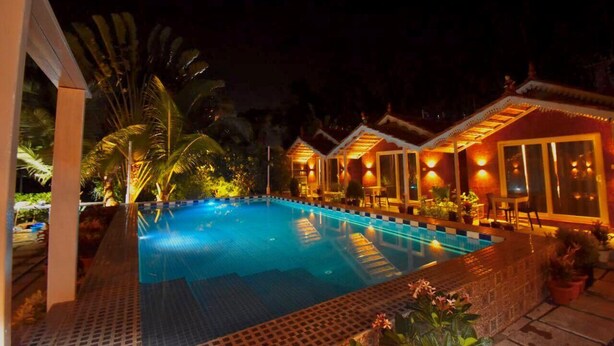 A swimming pool with a tiled deck, surrounded by potted plants and a building with large windows and warm lighting. - Golden Rice Resort (Candolim)