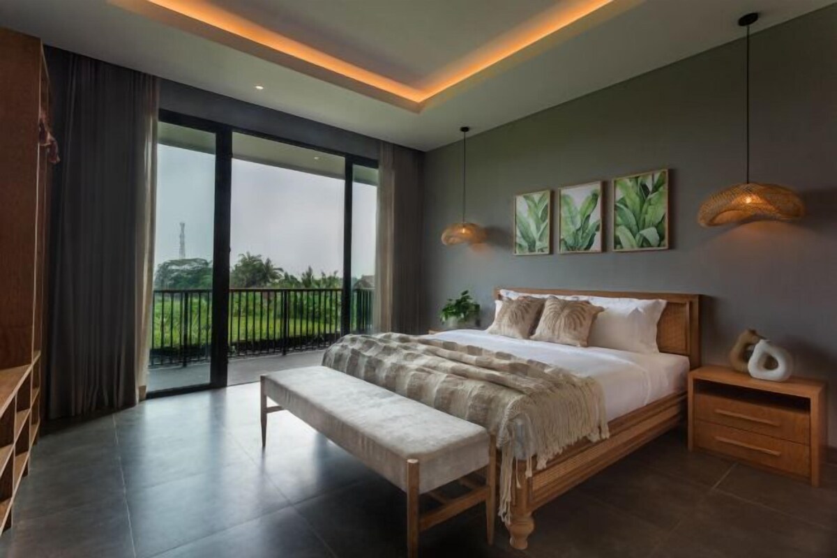 A modern bedroom with a large bed, a bench, and a view of greenery outside.