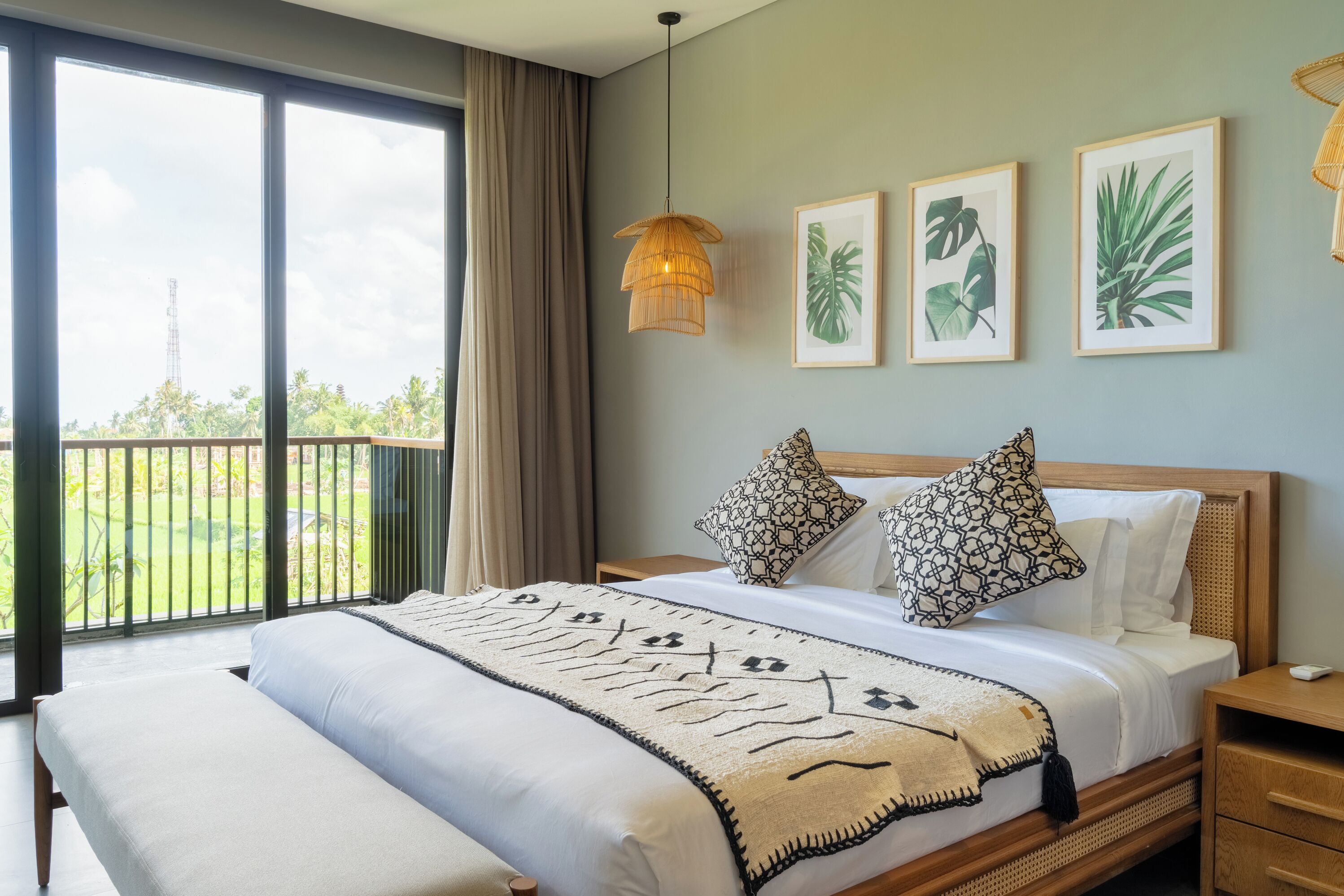 A bedroom with a large bed, two pillows, a bedside table, a nightstand, and a window with a view of greenery.