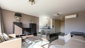 Een moderne woonkamer met een bank, een salontafel en een televisie. Er is een keuken met kasten en een eettafel.