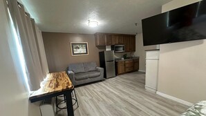 Een compact leefruimte met een kitchenette, een grijze bank, een houten eettafel en een wandgemonteerde flatscreen-tv.