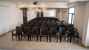 Une salle de conférence avec des rangées de chaises noires, des ventilateurs de plafond et une grande fenêtre.