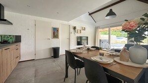 Una cocina moderna con comedor, que incluye una mesa de madera dispuesta para cuatro personas, sillas negras, un jarrón grande con flores y un televisor en la pared.
