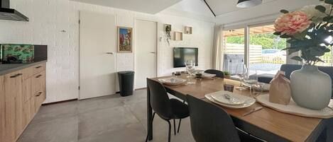Une cuisine moderne avec un coin repas, comprenant une table en bois dressée pour quatre personnes, des chaises noires, un grand vase rempli de fleurs et un téléviseur fixé au mur.