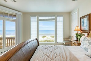 Een slaapkamer met een groot bed, een stoel, een bureau en uitzicht op het strand door grote ramen.