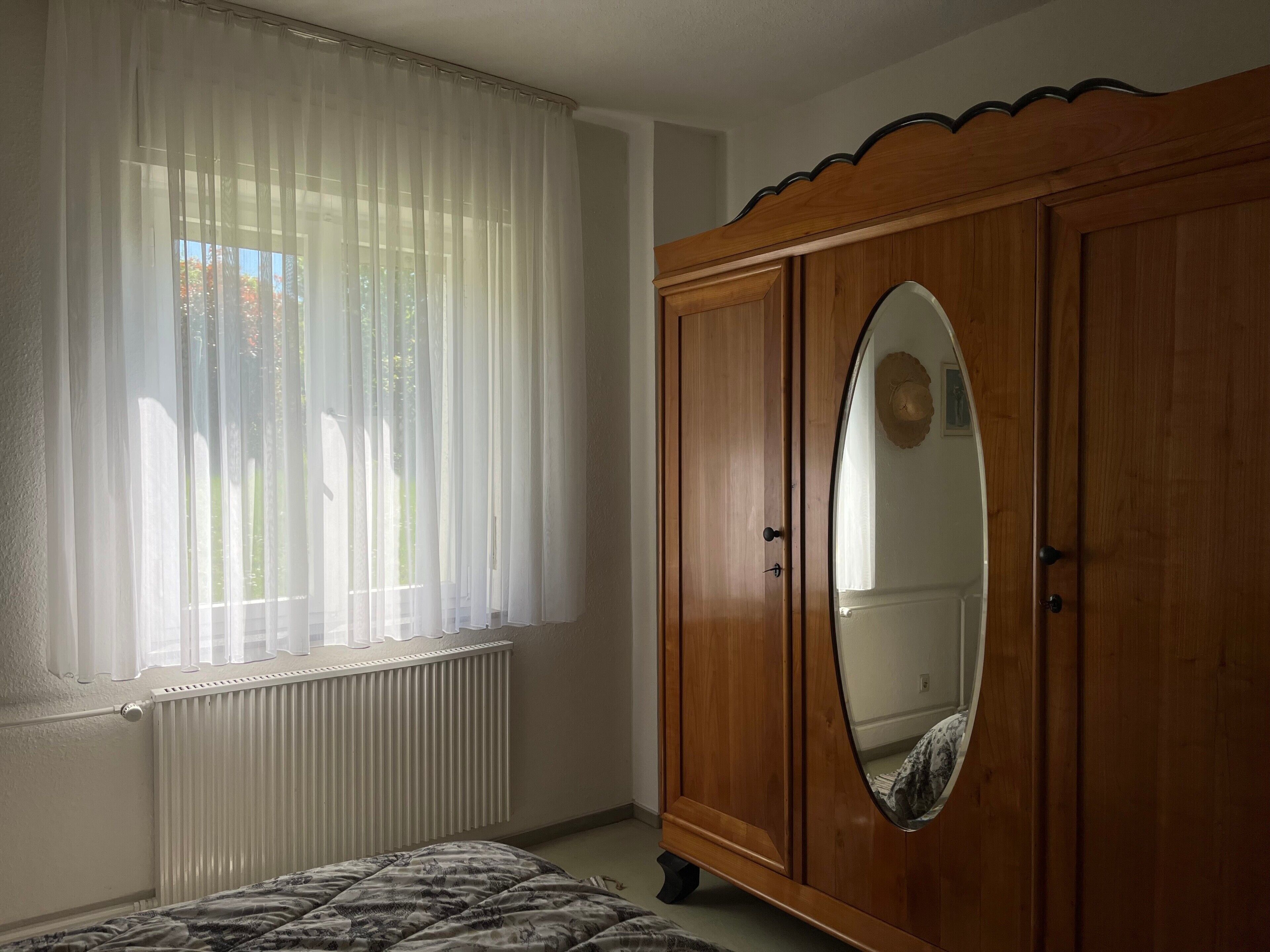 Een slaapkamer met een grote houten garderobe, een radiator en dunne gordijnen voor het raam.