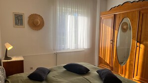 A bedroom with a bed, pillows, a nightstand with a lamp, a window with sheer curtains, a wooden wardrobe, and a framed picture on the wall. - Private Room 'Auf dem Karlsberg' with Wi-Fi (Weilburg)