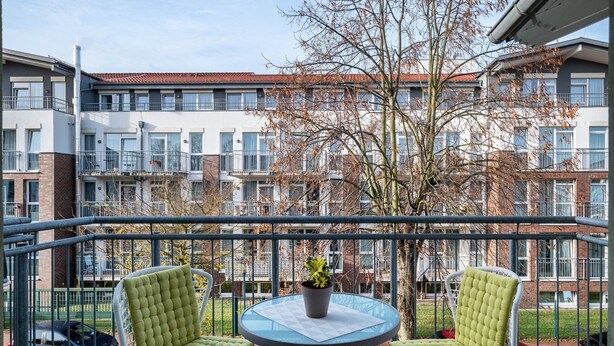 A balcony with two chairs and a table, a potted plant, and a view of a multi-story building with balconies. - Apartment 'Direkt Am' with Balcony and Wi-Fi (Graal-Müritz)
