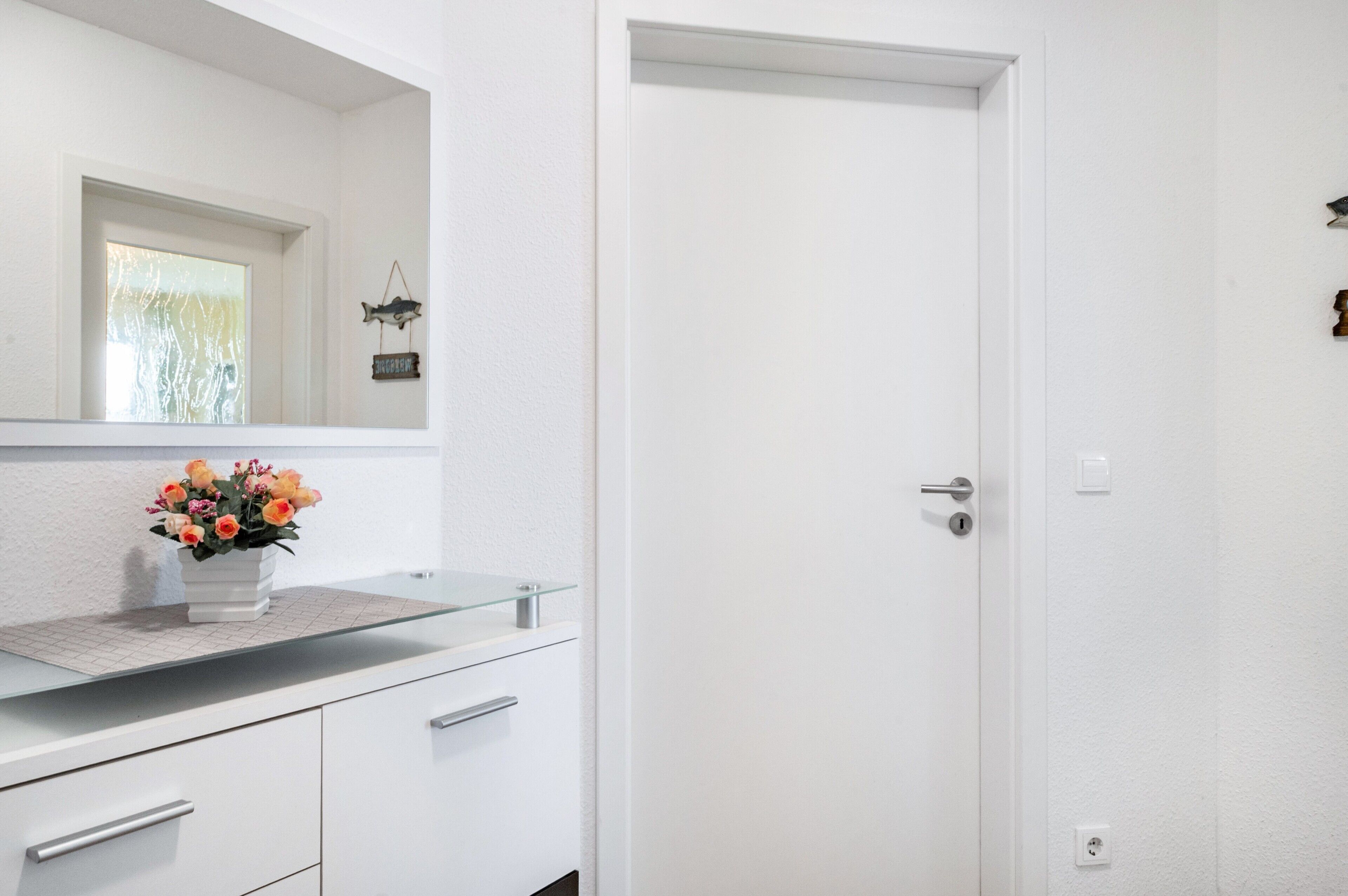 Un baño blanco con tocador de cristal, un espejo, una puerta y una ventana pequeña.