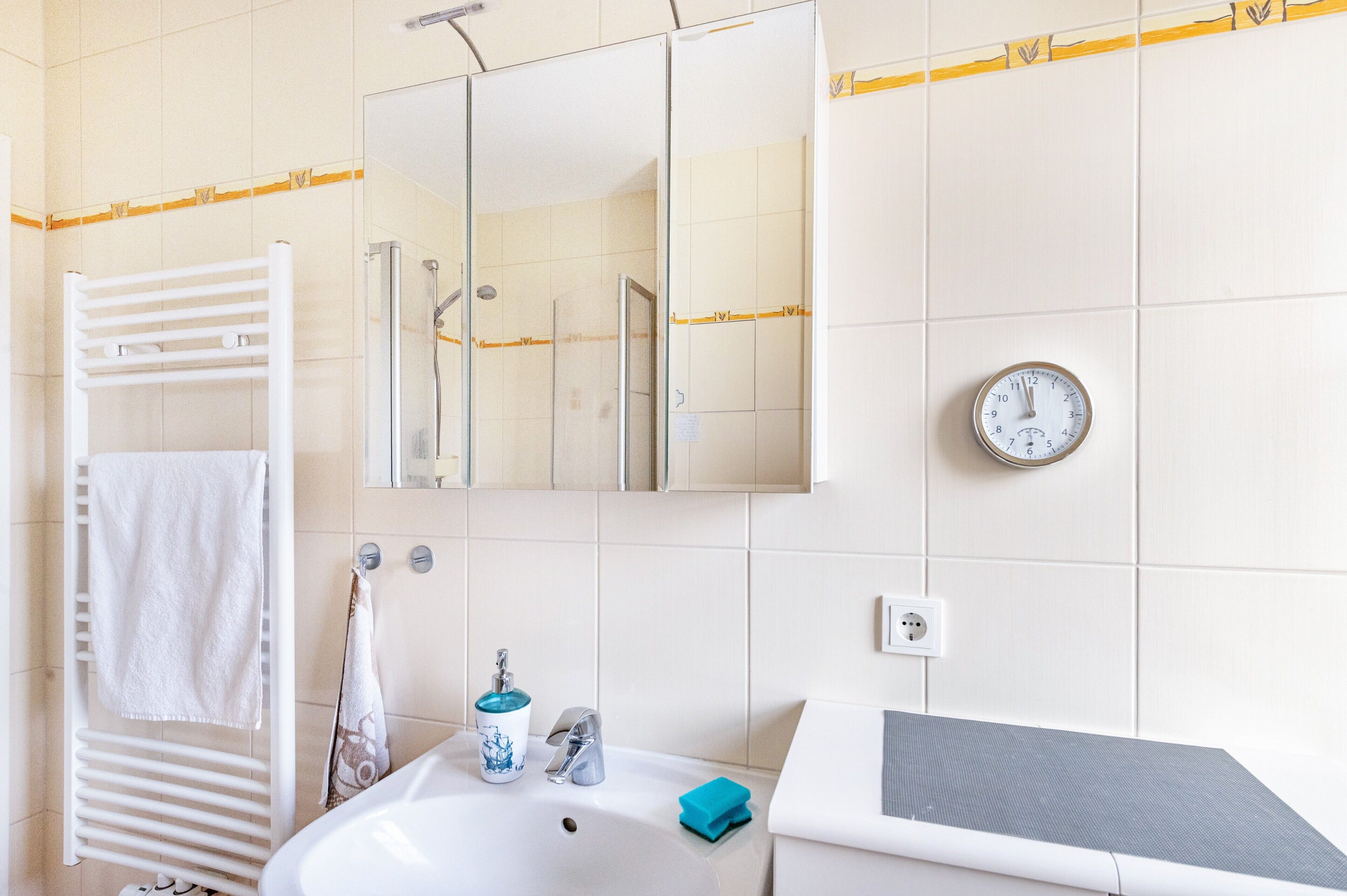 Un baño con lavamanos blanco, espejo, toallero y un reloj en la pared.