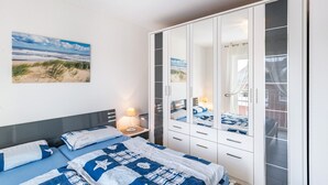 A bedroom with a bed, bedside table, lamp, wardrobe, and a beach view painting. - Apartment 'Direkt Am' with Balcony and Wi-Fi (Graal-Müritz)