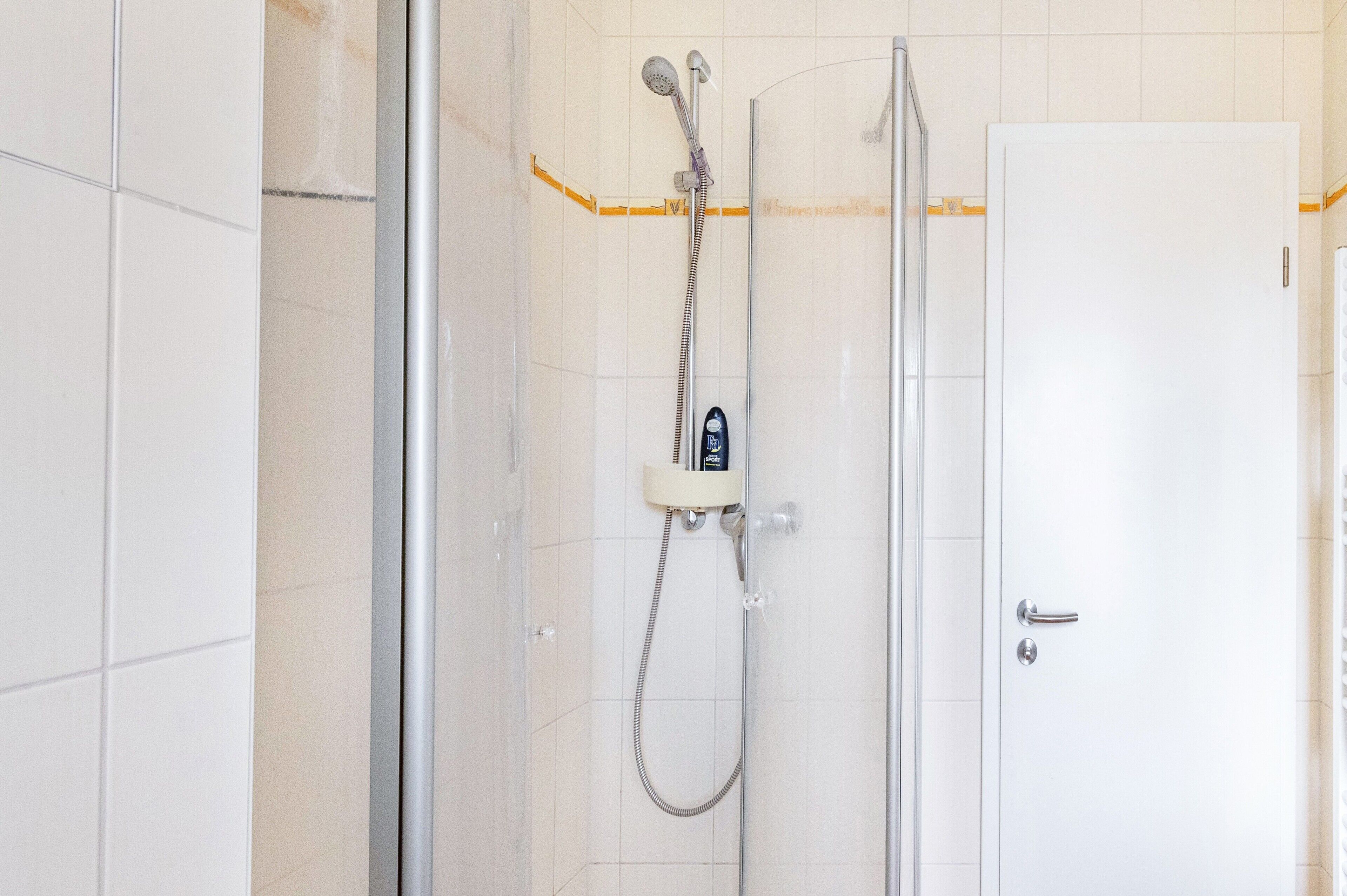 Ducha con cabezal de ducha manual, grifo empotrado en la pared, puerta de cristal y puerta blanca con manija plateada.