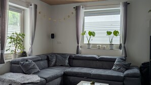 A modern living room with a sectional sofa, a coffee table, and a large window with curtains. - Apartment 'An Der Quell' with Private Terrace, Private Garden and Wi-Fi (Blankenheim)