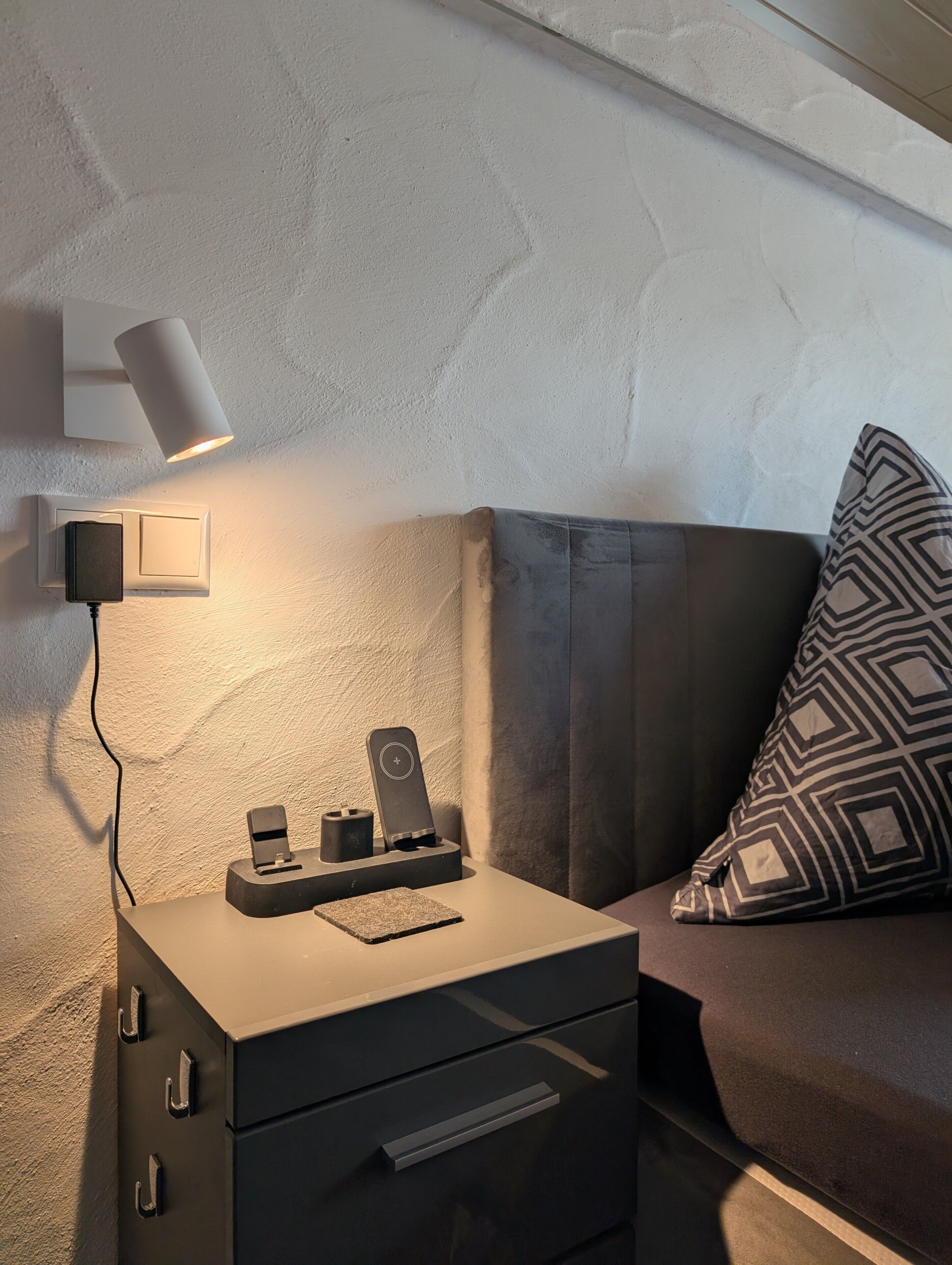 Una habitación moderna con mesita de noche, lámpara, enchufe y estación de carga.