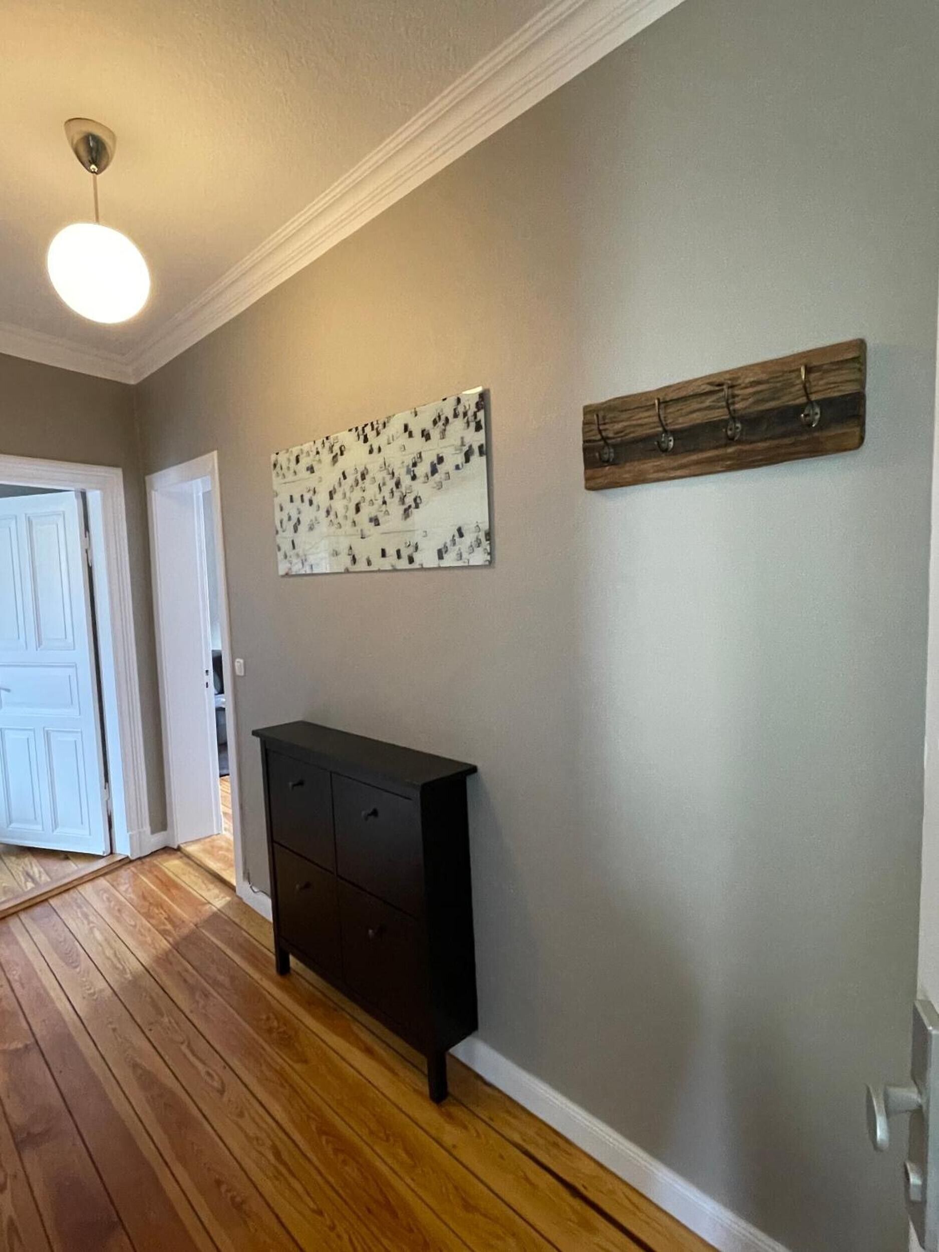 Ein Zimmer mit Holzboden, einer dunklen Kommode, einer Deckenleuchte, einer weißen Tür und einem an der Wand befestigten Kunstwerk.