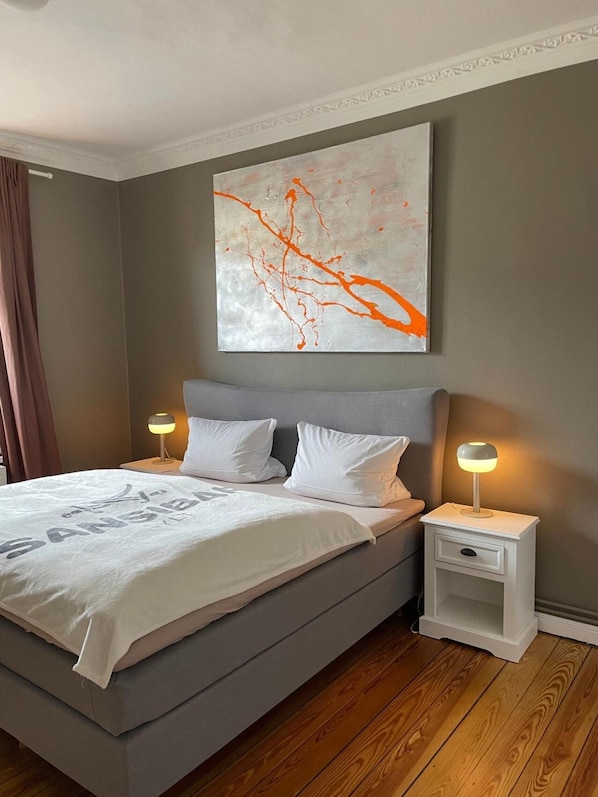 A modern bedroom with a grey bed, white pillows, a large abstract painting, and bedside lamps.