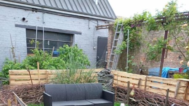 Une terrasse de jardin avec un canapé en osier noir, une petite table et une chaise. Un pont en bois entoure la terrasse, et un bâtiment en briques se trouve à l’arrière-plan.