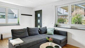 A modern living room with a grey sofa, a small table with a vase, and a window with a view of the outside. - Apartment 'Seeglück' with Mountain View, Private Terrace and Wi-Fi (Bodman-Ludwigshafen)