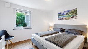 A bedroom with a bed, a desk, a chair, a window with a view of greenery, and a painting of a landscape. - Apartment 'Seeglück' with Mountain View, Private Terrace and Wi-Fi (Bodman-Ludwigshafen)