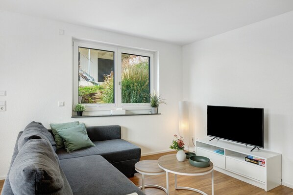 A modern living room with a sectional sofa, a flat-screen TV, a small round table, and a window with a view of greenery.