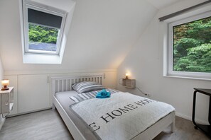 Une chambre avec un plafond en pente, un lit recouvert d’une courtepointe matelassée, une table de chevet avec une lampe, une fenêtre donnant sur la verdure et une étagère fixée au mur, également avec une lampe.