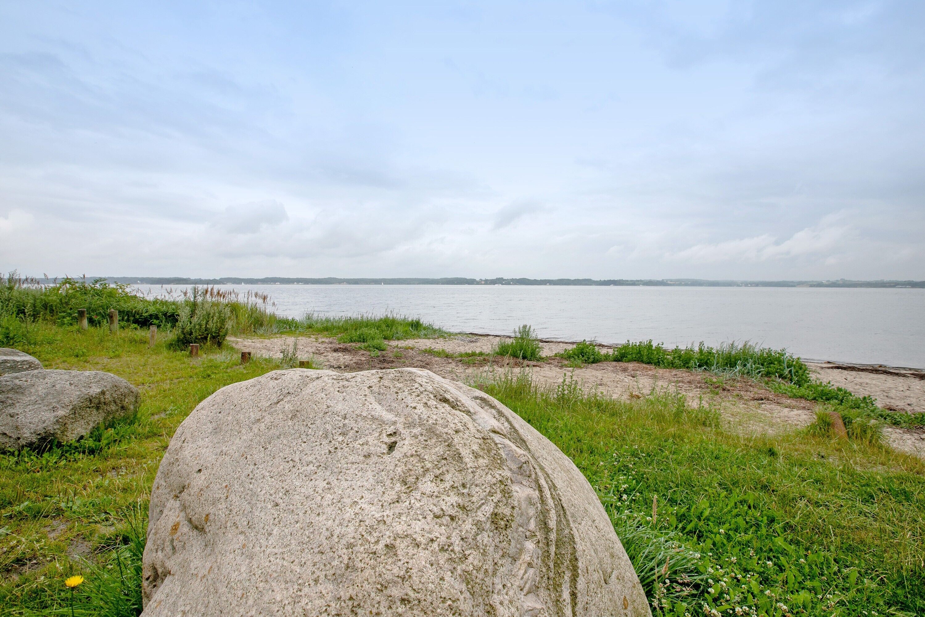 Een grote steen in een grasveld, met een waterpartij en een bewolkte lucht op de achtergrond.