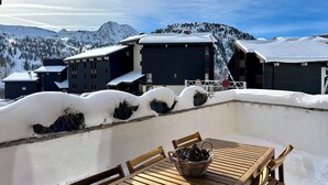 Une table en bois, des chaises et un panier, le tout placé devant un paysage montagneux enneigé avec des bâtiments.