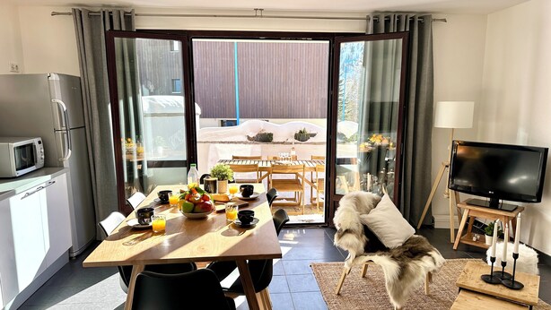 Un salon moderne avec un coin repas, une cuisine équipée d’un four à micro-ondes, un téléviseur posé sur un meuble et une vue sur l’extérieur.