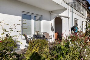 Un bâtiment blanc doté d’un balcon, une table avec des chaises et un jardin où poussent différentes plantes et fleurs.
