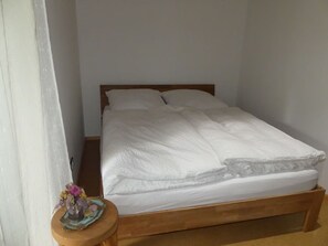 Un lit simple avec un cadre en bois, une literie blanche et une petite table sur laquelle est posé un vase de fleurs.